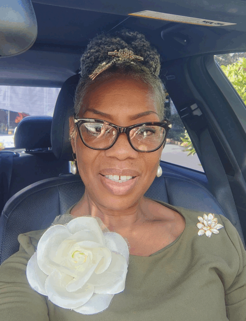 Smiling woman with glasses holding a white flower inside a car.